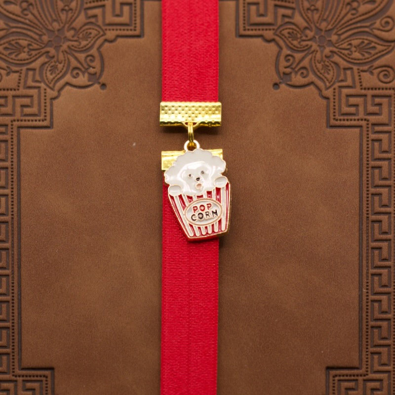Bookband elastic bookmark with a poodle dog in a popcorn bucket design on a red ribbon against a brown book